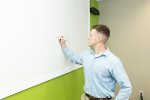 Man writes on white board against green wall.