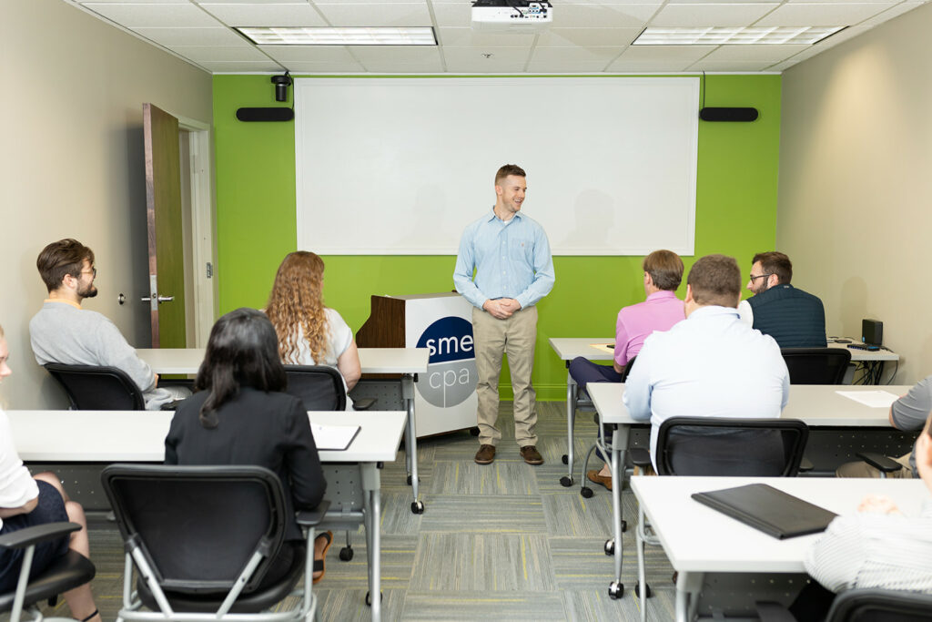 A man stands at the front of a conference room presenting to a group of people seated at tables.